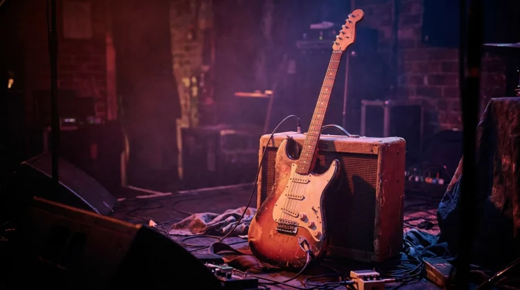 Guitarra eléctrica apoyada en un amplificador sobre un escenario, con iluminación de tonos rojos y morados